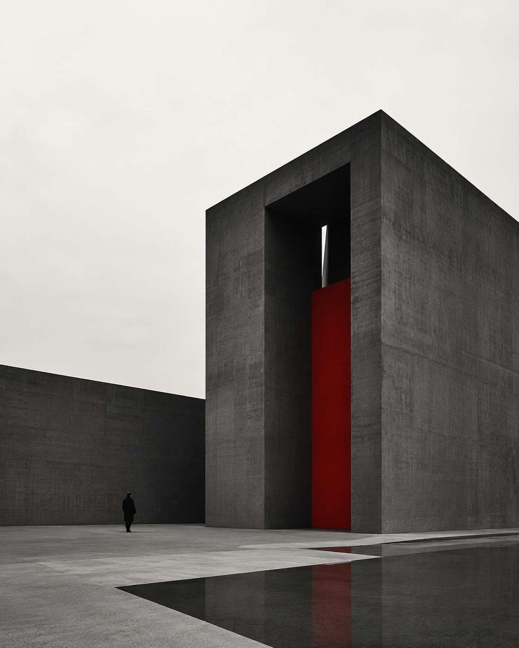 A monolithic concrete tower with a single vertical red slot, mirrored in a still reflecting pool.