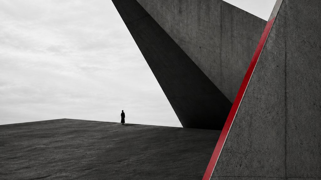 A concrete prow cantilevered over a dark plain, one red edge running down the wall beside it, a lone figure at the horizon.